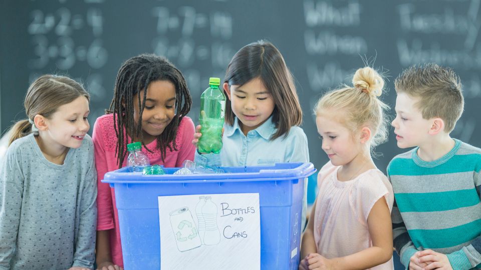 Five children surrounding a recycling bin and putting plastic bottles into it.